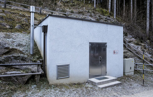 Auf dem Bild ist die Außenansicht eines teilweise in den Hang gebauten, kleinen Kraftwerkes, mit einem Eingang an der Vorderseite, zu sehen.