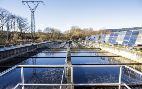 Auf dem Bild sind vertikal angeordnete, im Boden eingebrachte Wasserbehälter mit rechts an einer Wand fixierte PV-Module zu sehen.