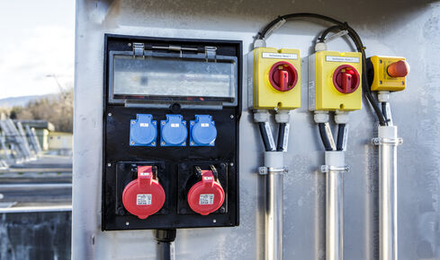 Auf dem Bild sind vertikal angeordnete, im Boden eingebrachte Wasserbehälter mit rechts an einer Wand fixierte PV-Module zu sehen.