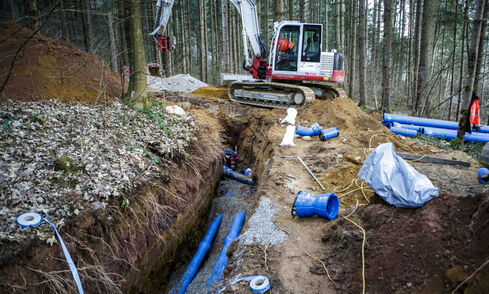 Auf dem Bild ist eine Baustelle in einem Waldstück mit offenem Graben, blauen Rohren und ein großer Bagger zu sehen.