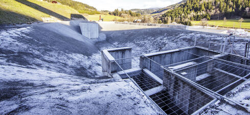 Auf dem Bild ist ein vertieftes, von hellem Kies und Schotter eingefasste Wasseransammlung mit zwei Betonbauten am oberen und unteren Ende zu sehen.