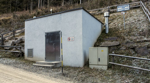 Auf dem Bild ist die Außenansicht eines teilweise in den Hang gebauten, kleinen Kraftwerkes, mit einem Eingang an der Vorderseite, zu sehen.