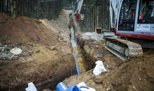 Auf dem Bild ist eine Baustelle in einem Waldstück mit offenem Graben, blauen Rohren und ein großer Bagger zu sehen.